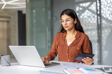 Young indian businesswoman in glasses calculating finances, managing budget, or performing accounting tasks using a laptop and calculator at her desk in a modern office