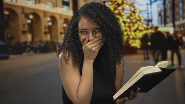 Young woman with glasses holding book on street with lights smiling with hand on mouth during evening