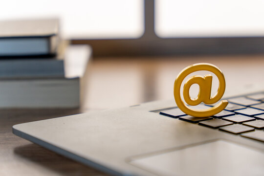 A yellow at sign placed on a laptop keyboard with stacked books in the background. Email, digital communication, internet, online services, and modern workspace concepts.