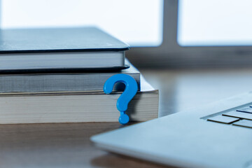 A small blue question mark placed between stacked books and a laptop. Inquiry, research, education, technical support, online study, confusion concepts.