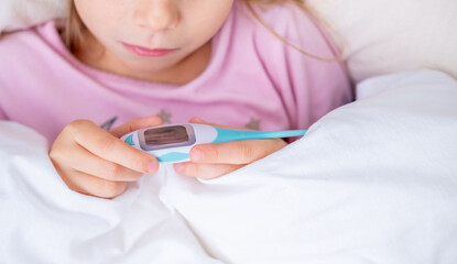 Little girl in pink pajamas under white blanket holds digital thermometer, checking temperature, Childhood illness and fever, home care and diagnosis, parental concern for health, sickness at home