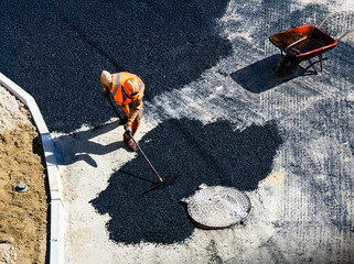 Un ouvrier de construction en uniforme orange et casque répand de l'asphalte avec un râteau sur...