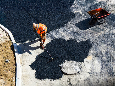Un ouvrier de construction en uniforme orange et casque r&eacute;pand de l'asphalte avec un r&acirc;teau sur une chauss&eacute;e, &agrave; c&ocirc;t&eacute; d'une brouette rouge