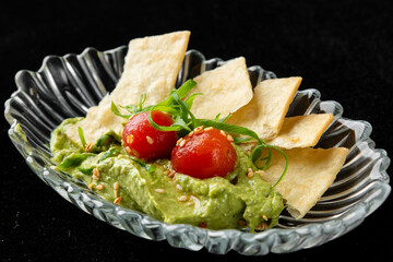 Appetizing close-up of fresh guacamole dip topped with cherry tomatoes, green onions, and sesame seeds, served in a glass dish with crispy flatbread crackers on a black background.