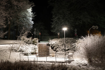 Snow-covered stairs, benches, and frosty trees illuminated by streetlights on a quiet winter night in a peaceful park.