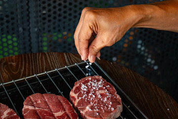 Chef hand seasoning raw ribeye steak cuts with coarse sea salt on a stainless steel tray