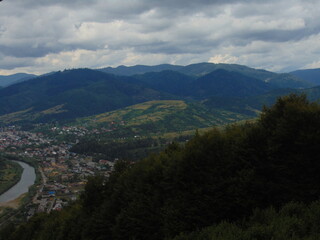 A peaceful mountain valley with rolling green hills, a winding river, and a small town nestled below. Dramatic clouds cast soft shadows, creating a calm and scenic landscape