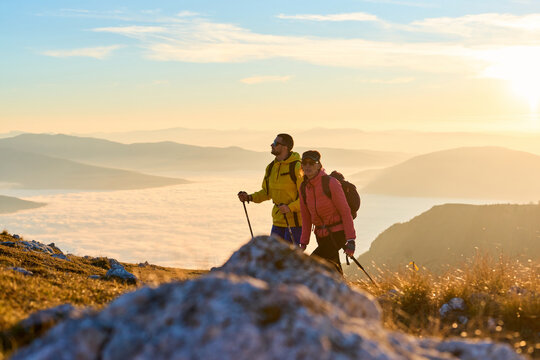 Couple hiking through a grassy mountain trail during sunrise, surrounded by a stunning backdrop of fog in the valleys below. The warm light creates a beautiful atmosphere