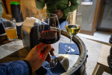 Close-up of a person holding a glass of red wine at a rustic table with drinks. Two wine glasses and a beer glass are placed on a wooden barrel, creating a relaxed evening atmosphere.