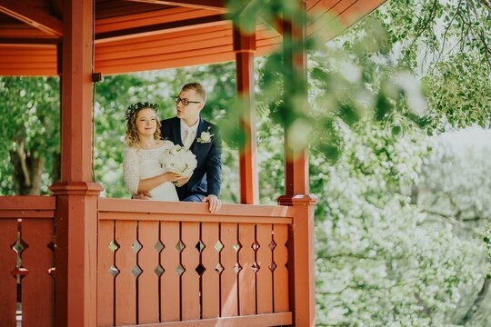 Bride and groom standing together on a wooden gazebo, surrounded by lush greenery, celebrating their love in a romantic outdoor wedding setting with natural light