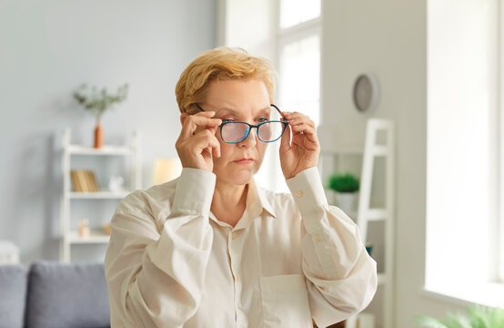 Portrait of focused woman adjusting her glasses while standing in bright room. Senior lady carefully positioning glasses on her face with both hands and looking intently at something in front of her.