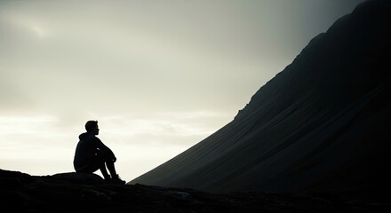 A solitary man in silhouette sits on a rocky outcrop, contemplating the vast and moody landscape with a dramatic mountain slope under a pale, overcast sky