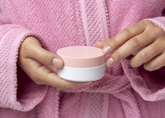 Closeup of woman in pink bathrobe holding cream jar with pink lid in hands, cosmetic mockup