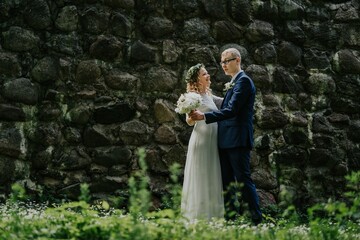 Couple in wedding attire stands joyfully in a lush green field, surrounded by wildflowers, with a rustic stone wall in the background, celebrating their love and commitment