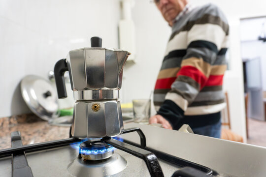 Metal stovetop coffee maker boiling on a gas burner while an older man stands in the blurred background, capturing a warm and authentic moment of home kitchen life.