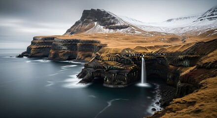 Majestic long exposure landscape of a powerful waterfall flowing from a coastal cliff into the sea, with a snowy mountain range under a moody, overcast sky