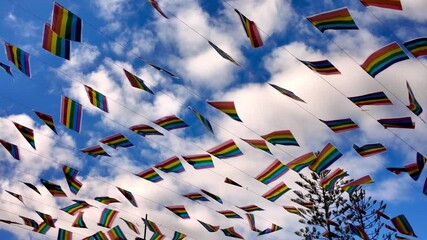 Low angle view of many rainbow LGBT pride flags waving in slow motion against a beautiful blue sky with white clouds. Pride month celebration, gay freedom, and lgbtqia plus rights concept