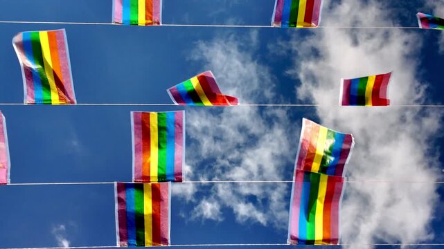 Colorful rainbow flags hanging on strings waving in the wind against a beautiful blue sky with white clouds, symbolizing the gay pride movement, freedom, and equality for all people