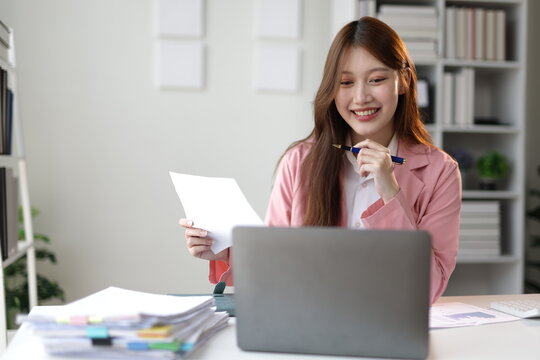 Young professional woman smiling while reviewing printed documents and comparing them with data displayed on her laptop screen.
