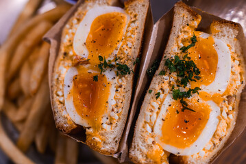 A close up of a Japanese tamago sando in Tokyo shows creamy egg salad, halved soft boiled eggs, chopped herbs, and pepper in a paper sleeve beside golden fries.