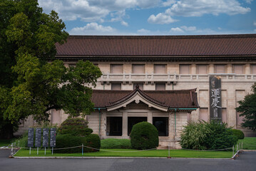 Obraz premium Visitors arrive at Tokyo National Museum in Ueno Park. Stone facade, tiled roof, gabled entry, vertical Japanese sign, cordoned path, soft daylight, formal mood.