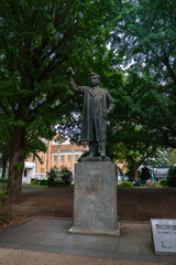 Obraz premium A bronze man in a long coat raises an arm on a tall pedestal in a Tokyo campus park, brick academic buildings through trees and a signboard near the monument.