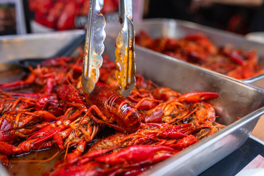 Close up of a vendor lifting a glossy crawfish from a simmering tray at a Tokyo market during meal rush, with steam, oily broth, and casual serving setup visible.