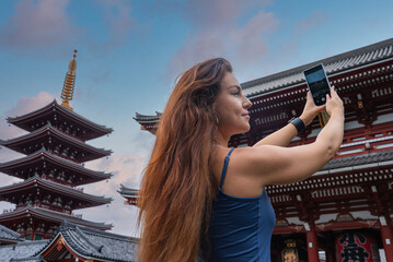A traveler holds a smartphone for a selfie at Senso ji in Asakusa, Tokyo. The five story pagoda and Kaminarimon style details are visible in daylight.