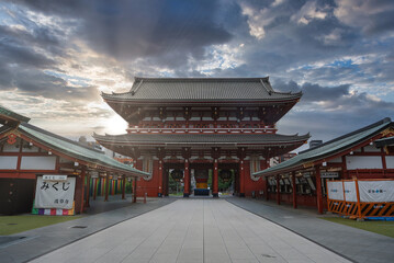 Obraz premium A broad stone path leads to Hozomon Gate at Senso ji Temple in Asakusa, Tokyo, with red and white stalls, omikuji signs, and tiered roofs in soft early light.