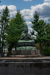 Obraz premium A bronze Buddha sits in prayer on a lotus pedestal in a Tokyo temple courtyard, framed by tall trees and garden stones, captured during daytime urban travel exploration.
