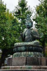 Fototapeta premium A bronze Buddha sits on a lotus pedestal in Tokyo, framed by tall ginkgo trees. Ritual objects and an ornate headdress show temple craft at Asakusa or Ueno.