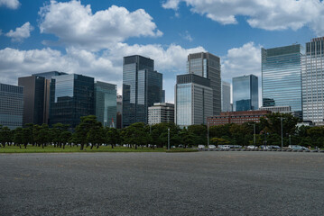 Gravel forecourt leads to pruned pines and Tokyo Marunouchi towers, with red brick buildings, parked cars, and light poles near Imperial Palace grounds in daylight. © Aerial Film Studio