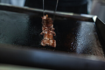 Close up shows a vendor searing skewered meat on a teppan griddle in Tokyo, turning with metal...