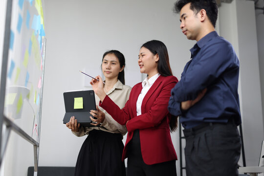 Asian businesswoman leading meeting with sticky notes on whiteboard and digital tablet.