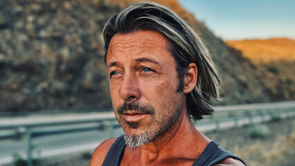 Middle aged tanned man with half long blond hair and grey stubble beard in singlet shirt walking outdoors during golden hour. Hills of Almogia, Malaga, Andalusia, Spain.