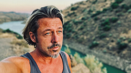 Middle aged tanned man with half long blond hair and grey stubble beard in singlet shirt walking outdoors during golden hour. Hills of Almogia, Malaga, Andalusia, Spain.