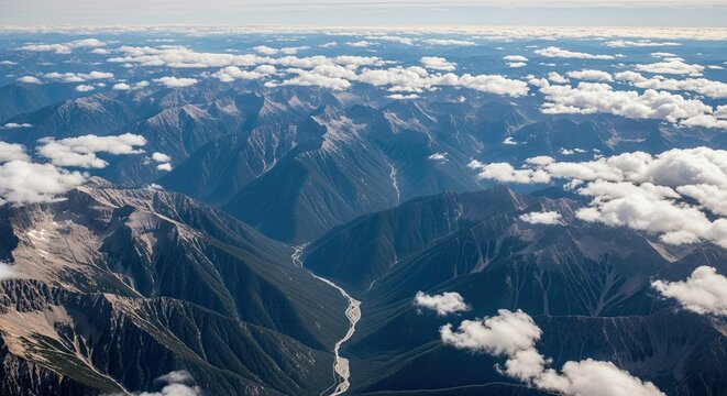 Majestic aerial panorama of a rugged mountain range with a winding river in a deep valley, viewed from high altitude above fluffy white clouds under a blue sky - Powered by Adobe