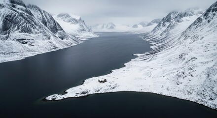 Dramatic aerial view of a solitary wooden cabin on a snowy peninsula surrounded by a dark fjord and majestic, snow-covered mountains under an overcast sky