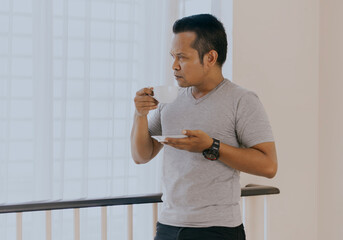 Young asian man with cup of coffee looking in window at home interior.