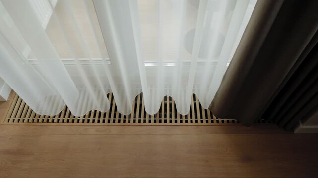In-floor convector heating system with linear wooden grille in a refined modern living space illuminated by soft daylight and airy white curtains