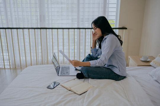 Focused asian woman works on financial documents, sorting reports and analyzing data at home, sitting on bed in cozy bedroom interior. Accounting and finance concept.