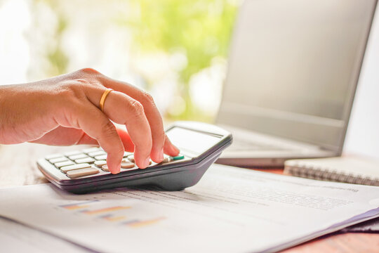 Selective focus on woman’s hand using a calculator with financial documents in front of laptop, remote work concept