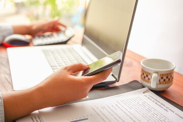 Woman&rsquo;s hand holding a smartphone in front of laptop on wooden desk with documents, remote work concept