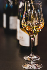 A close-up shot of a stemmed tasting glass filled with amber liquid, possibly white wine or whiskey, on a dark table with blurred bottles in the background