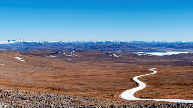 A winding road traverses a desolate, rocky, and brown tundra landscape, leading towards a distant lake and snow-capped mountains under a bright blue sky.