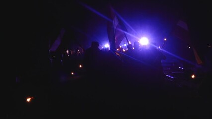 Silhouettes of people holding Latvian flags illuminated by purple stage light at night, surrounded by torches during a solemn gathering or commemoration.
