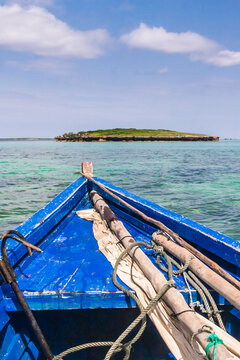 A traditional wooden boat with blue painted planks and a removable mast and furled sail, an island in the distance. 