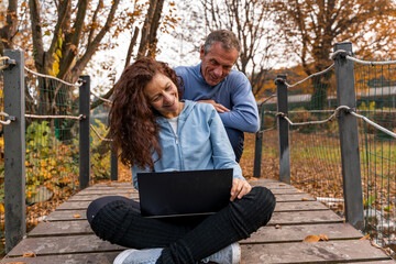 Happy senior couple sharing a laptop together on a wooden bridge in an autumn park