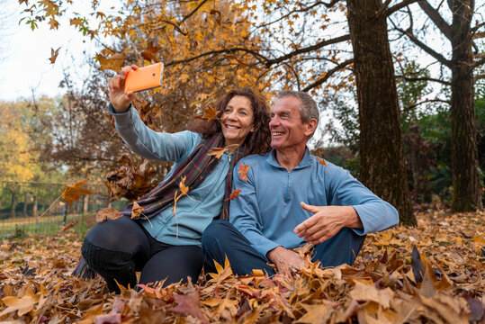 Happy senior couple sitting in fallen autumn leaves, smiling and taking a selfie - Powered by Adobe