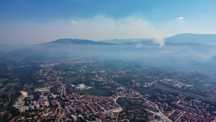 Elevated view of a densely populated area under a blanket of smoke/mist, with a faint column of smoke rising over the distant hills